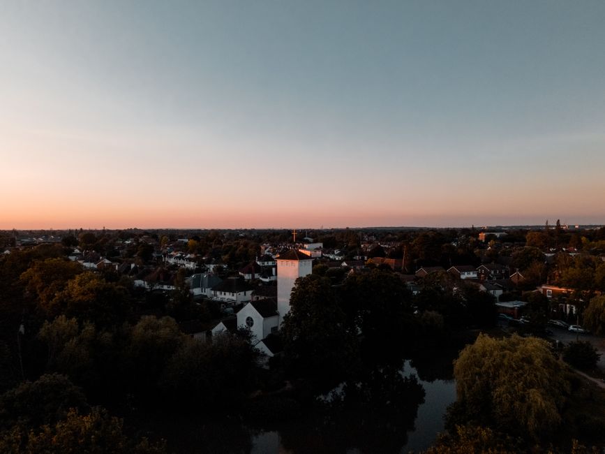 An aerial view of a suburban residential area during sunset, featuring tightly packed houses with white facades and dark roofs, interspersed with numerous large trees with green foliage. In the foreground, trees partially obscure the rooftops, while a white church with a tall spire stands prominently near the center of the image. The sky above displays a gradient of colors transitioning from soft orange near the horizon to pale blue higher up, casting a gentle light over the scene. The landscape extends into the distance with a flat horizon, and no vehicles or people are visible in the image. This scene exemplifies a quiet neighbourhood suitable for private waste collection or on-site clearance, aligning with the services of wastecollectionsurrey.co.uk for effective rubbish removal in Surrey. The subdued natural lighting highlights environmental calmness, and the composition emphasizes the residential setting, supporting alternative waste handling options for local residents or service providers such as Waste Collection Surrey.