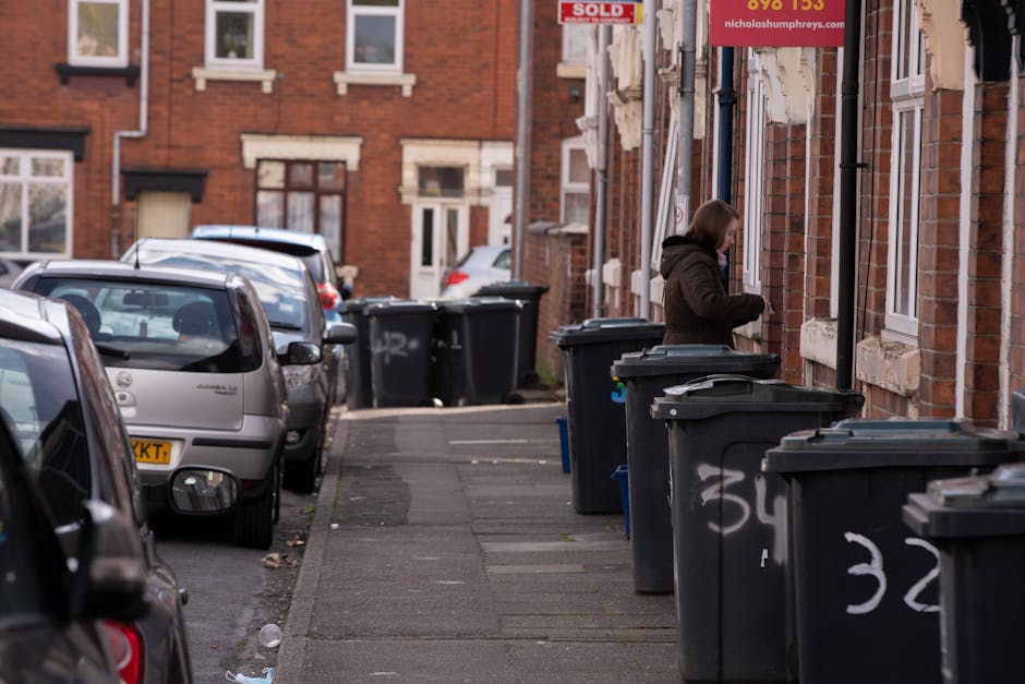 A woman standing on a narrow city sidewalk next to a row of black wheelie rubbish bins with closed lids, positioned along a brick building frontage. The bins have white painted numbering on them and are placed on a concrete pavement with slight wear and scattered litter. Behind the woman, there are multiple small, traditional brick houses with white window frames and some with partial brick arch detailing above windows. To the left, several parked cars, including a silver hatchback, line the street, with additional vehicles visible further along the road. The scene appears to be during daylight hours with soft, natural lighting. The setting suggests a typical urban residential area, possibly accommodating private waste collection and alternative rubbish disposal methods, as indicated by the arrangement of the bins outside individual dwellings. Waste Collection Surrey's presence is implied through the context of rubbish management, although not directly visible in the image.