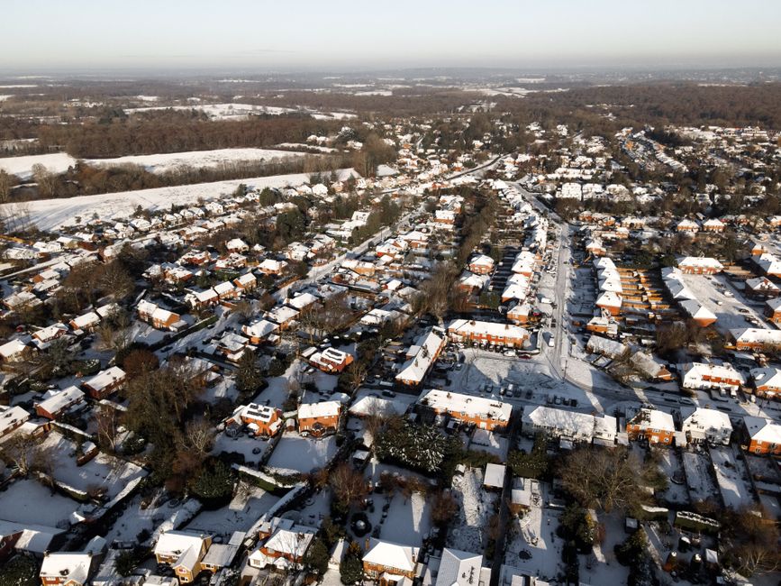 An aerial view of a suburban residential area during sunset, featuring tightly packed houses with white facades and dark roofs, interspersed with numerous large trees with green foliage. In the foreground, trees partially obscure the rooftops, while a white church with a tall spire stands prominently near the center of the image. The sky above displays a gradient of colors transitioning from soft orange near the horizon to pale blue higher up, casting a gentle light over the scene. The landscape extends into the distance with a flat horizon, and no vehicles or people are visible in the image. This scene exemplifies a quiet neighbourhood suitable for private waste collection or on-site clearance, aligning with the services of wastecollectionsurrey.co.uk for effective rubbish removal in Surrey. The subdued natural lighting highlights environmental calmness, and the composition emphasizes the residential setting, supporting alternative waste handling options for local residents or service providers such as Waste Collection Surrey.