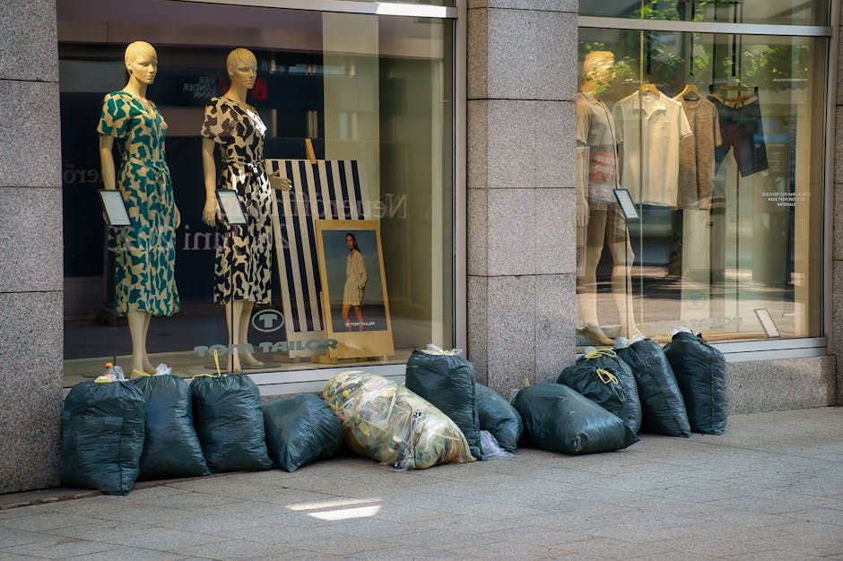 A row of dark green garbage bags filled with waste is lined up along the pavement outside a clothing store. The bags are tied at the top, with some showing slight bulges indicating their contents, and one bag appears to be partially transparent, revealing discarded paper and plastic inside. These bags are positioned directly in front of a large glass window display, which showcases mannequins dressed in colorful summer outfits and various clothing items hanging on racks inside the store. The store's exterior features stone cladding framing the window, with sunlight casting soft shadows on the pavement. The scene suggests an instance of waste awaiting collection, possibly as part of an independent rubbish removal service such as those offered by Waste Collection Surrey, suitable for retail or commercial premises seeking private waste management outside of standard council services.