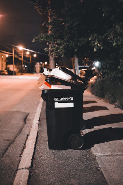A black wheeled rubbish bin with a white label and the text 'ST. JOHN'S' on its front, positioned on a sidewalk beside a curb in a residential area at night. The bin is overflowing with a mixture of discarded paper, cardboard, and plastic waste. Nearby, other bins are visible in the background, some partially open and filled with waste materials. The scene is illuminated by streetlights casting a warm, orange glow, with shadows stretching across the pavement and nearby trees and bushes lining the sidewalk. The surrounding environment suggests an urban or suburban setting with quiet streets, and the image relates to waste collection or rubbish removal services, possibly indicating an alternative or private waste management context often handled by companies such as Waste Collection Surrey, especially in relation to on-site clearance or tailored rubbish disposal solutions.
